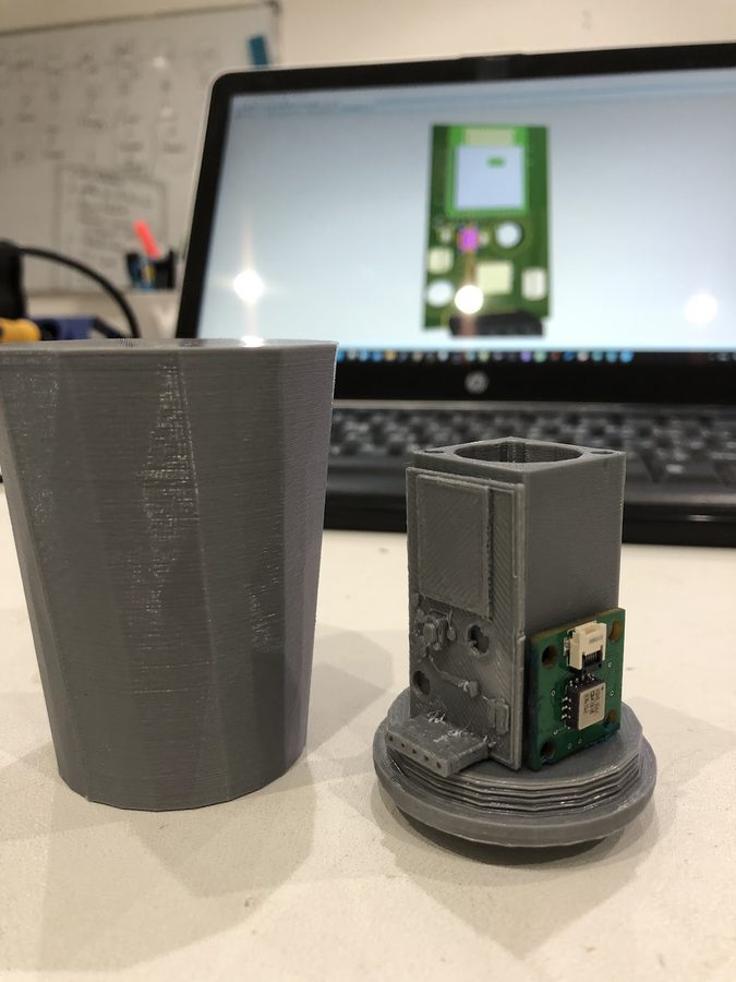 3D-printed grey sensor enclosure prototypes next to a laptop showing the PCB design
